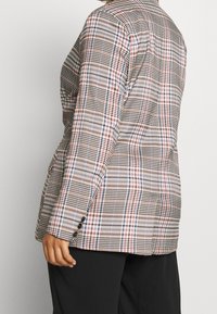 Osoba ubrana w dopasowaną marynarkę w kratę w czerwone, niebieskie i czarne linie, w połączeniu z czarnymi spodniami, pokazana od połowy torsu do górnej części uda.