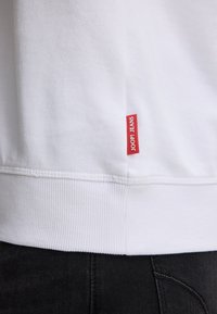 Sweatshirt bianco in cotone con orlo a coste, completo di un piccolo etichetta rossa "JOOP! JEANS" sul lato, abbinato a pantaloni in denim scuro.