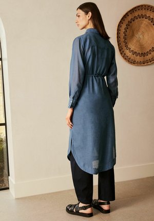 Langes, hellblaues Hemdkleid aus weichem Stoff, mit Kragen, langen Ärmeln, einem gebundenen Taillengürtel und einem abgerundeten Saum, kombiniert mit schwarzen Hosen und Sandalen.
