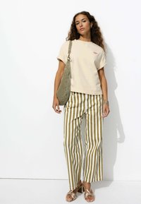 T-shirt en coton beige à manches roulées, pantalons à larges rayures vert olive, et sandales à talons métalliques. Sac fourre-tout en tissu doux.