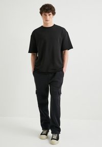 Schwarzes, übergroßes T-Shirt mit kurzen Ärmeln, kombiniert mit schwarzen Cargo-Hosen mit Seitentaschen und schwarzen Turnschuhen mit cremefarbenen Sohlen.