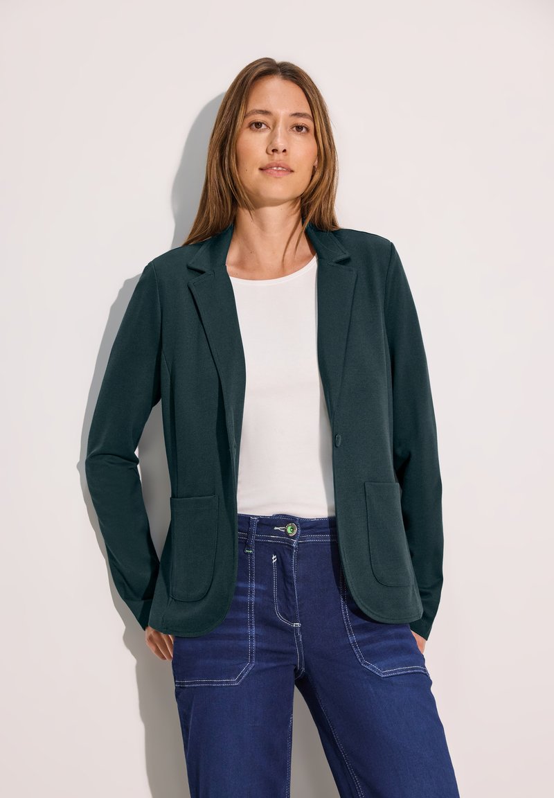 Blazer ajustado de color verde oscuro con dos bolsillos frontales y cierre de botón único. Combinado con una blusa blanca y jeans azul oscuro, mostrando un aspecto personalizado.