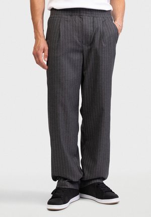 Άνδρας που φορά γκρι σκούρο παντελόνι με λεπτές ρίγες και μαύρα αθλητικά, στέκεται με το ένα χέρι στην τσέπη, μπροστά σε ένα απλό φόντο.