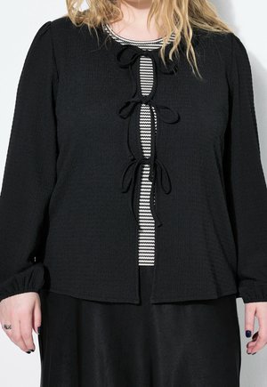 Femme portant un cardigan noir texturé noué avec trois nœuds sur un haut rayé noir et blanc et une jupe noire.