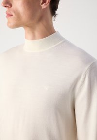 Maglione color crema con colletto alto a costine, texture morbida e discreto logo CK sul petto. Design aderente, minimalismo nei dettagli.