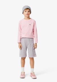 Camicia a maniche lunghe a righe rosa e bianche con un logo verde, abbinata a pantaloni corti grigi e sneakers rosa. Berretto grigio in lana indossato sulla testa.