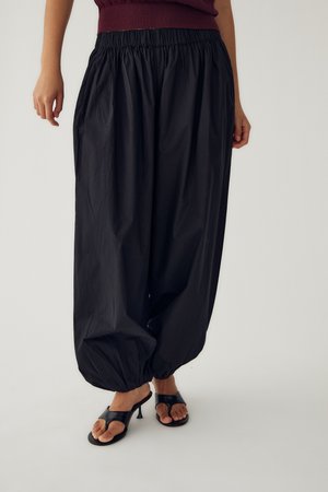 Losse zwarte broek met geplooide elastische manchetten, gedragen met zwarte open-toe sandaal met hakken op een persoon die staat tegen een effen achtergrond.
