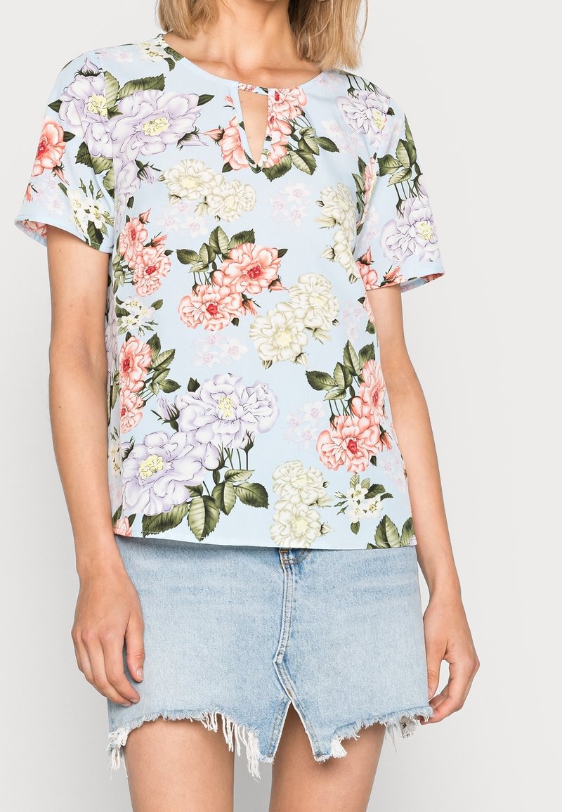 Femme portant un haut à manches courtes bleu clair à motifs floraux avec un col clé, associé à une mini-jupe en jean à ourlet effiloché.