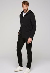 Giacca nera con cappuccio, due tasche frontali, chiusura con bottoni e maniche lunghe; abbinata a pantaloni neri e sneakers bianche su uno sfondo neutro.
