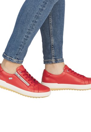 Personne portant un jean bleu et des baskets en cuir rouge avec des semelles blanches et un détail de fermeture éclair sur le côté, pieds croisés aux chevilles.