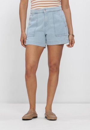 Vrouw draagt lichtblauwe denim short met grote voorzakken, een gestreept topje en beige instap loafers, staand op een effen achtergrond.