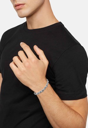 Bracelet en chaîne argentée avec des nœuds entrelacés, porté sur une main contre un fond de t-shirt noir. Texture lisse et finition métallique.