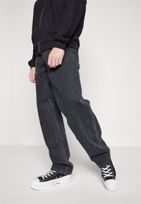 Sudadera negra y pantalones de mezclilla oscuros y sueltos, con costuras blancas y bolsillos laterales, combinados con zapatillas negras y blancas.