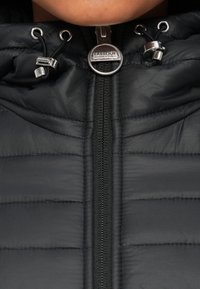 Μαύρο μπουφάν τύπου puffer με οριζόντιο καπιτονέ σχέδιο. Διαθέτει ασημένιο φερμουάρ με στρογγυλό μεταλλικό λογότυπο και ρυθμιζόμενα κορδόνια στο καπό.