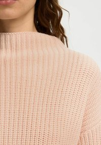 Svijetloružičasti pleteni pulover s rebrastom teksturom i visokim ovratnikom, obložen opuštenim krojem i mekanim, glatkim predivom.