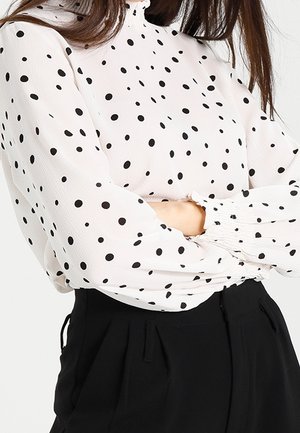 Blouse blanche à pois noirs, col montant et poignets élastiqués. Portée avec un pantalon noir au tissu texturé.
