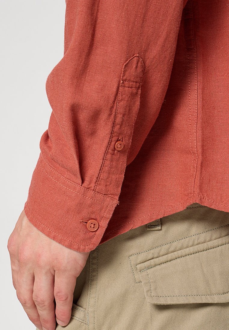 Chemise à manches longues couleur rouille en tissu texturé, avec des poignets à boutons et une finition lisse. Portée avec un pantalon clair.