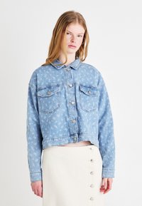Svetlo modra kratica jeans jakna z cvetličnim vzorcem, dvema žepoma na prsih in srebrnimi gumbi. Nosi se preko krila v kremni barvi z gumbi spredaj.