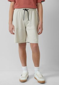 Beige Sportshorts mit elastischem Bund und schwarzem Kordelzug, ausgestattet mit zwei seitlichen Taschen und einer glatten, weichen Textur.