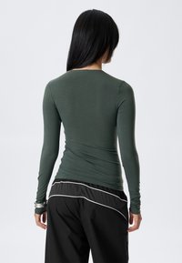 Long-sleeve, fitted top in dark green fabric with a smooth texture. Features a round neckline and is paired with loose black pants with white accents.