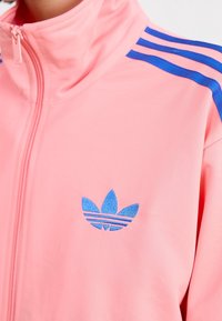 Rosa zip-up träjacka med blå ränder på axlarna och en blå broderad logotyp med tre blad. Slät textur, atletisk design.