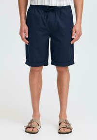Marineblaue Baumwollshorts mit lockerer Passform, elastischem Bund, Kordelzug, hochgerollten Säumen und Seitentaschen, kombiniert mit braunen Sandalen.