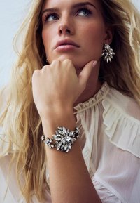 Srebrna narukvica s cvjetnim uzorkom koja sadrži velike, prozirne, okrugle i marquise rezane rhinestone kamenčiće. Model nosi usklađene naušnice; mekana, lepršava bluzica.
