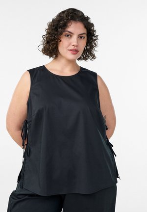 Kvinde med krøllet mørkt hår iført en ærmeløs sort top med siderem, stående foran en ensfarvet hvid baggrund.