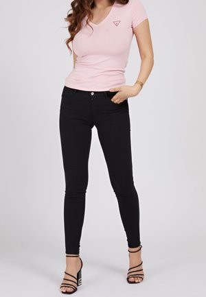 Kobieta ubrana w dopasowany jasnoróżowy t-shirt z dekoltem w serek, czarne obcisłe jeansy i czarne sandały na wysokim obcasie z paskami, stojąca z jedną ręką w kieszeni.