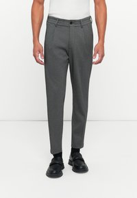 Uomo che indossa pantaloni slim-fit grigio scuro, calzini neri e scarpe slip-on nere, in piedi davanti a uno sfondo bianco uniforme.