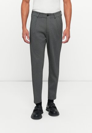 Man met slim-fit donkergrijze broek, zwarte sokken en zwarte instappers, staande tegen een effen witte achtergrond.