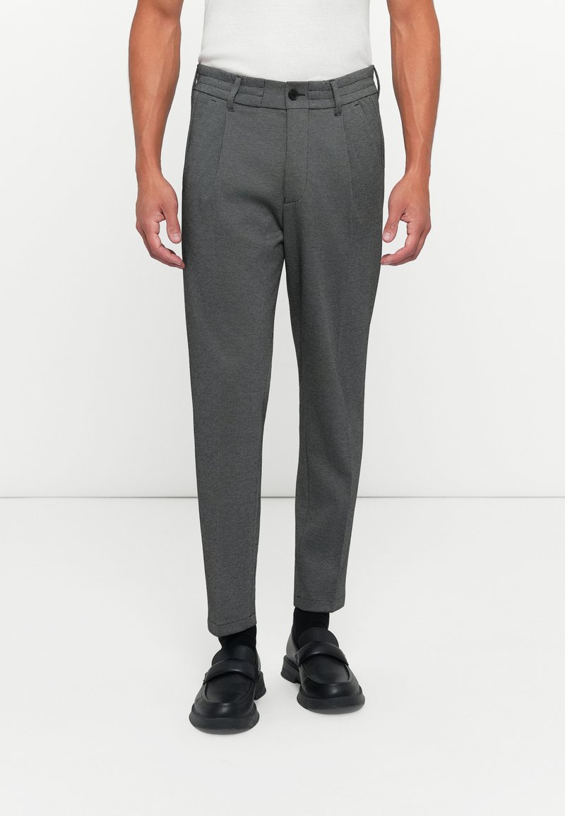 Uomo che indossa pantaloni slim-fit grigio scuro, calzini neri e scarpe slip-on nere, in piedi davanti a uno sfondo bianco uniforme.