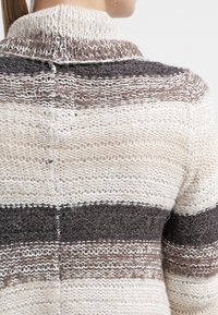 Persona vestida con un suéter de punto con anchas franjas horizontales en crema, marrón y gris oscuro, mostrado desde la parte de atrás.