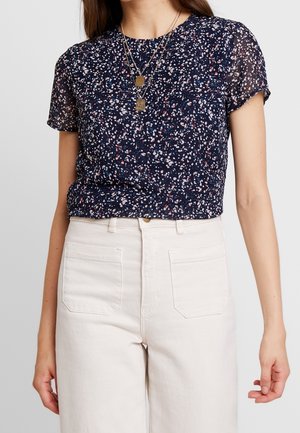 Kobieta w granatowej bluzce z krótkim rękawem z białymi i różowymi kropkami, dwoma złotymi naszyjnikami i wysokim stanem białych spodni z przednimi kieszeniami.