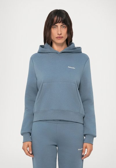 Woman wearing matching blue hoodie and sweatpants with small white text logos, standing against a plain light background.