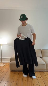 Young man in white shirt and green cap holding black long-sleeve shirt on hanger in a living room with wooden floor.