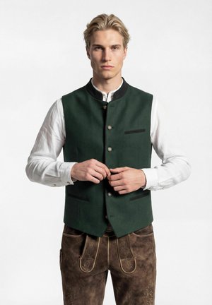 Junger Mann, der Knöpfe an einer grünen traditionellen Weste schließt, trägt ein weißes Hemd und braune bestickte Lederhosen vor einfachem Hintergrund.