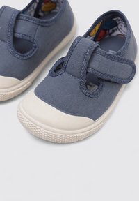 Zapatillas de lona azules con una correa de velcro, punta redonda y suela de goma blanca con patrones texturizados y un forro interior colorido.