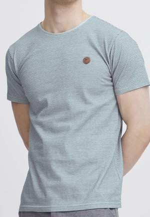 Mann trägt ein hellblau-weiß dünn gestreiftes kurzärmeliges T-Shirt mit kleinem rundem braunen Logo auf der linken Brust.