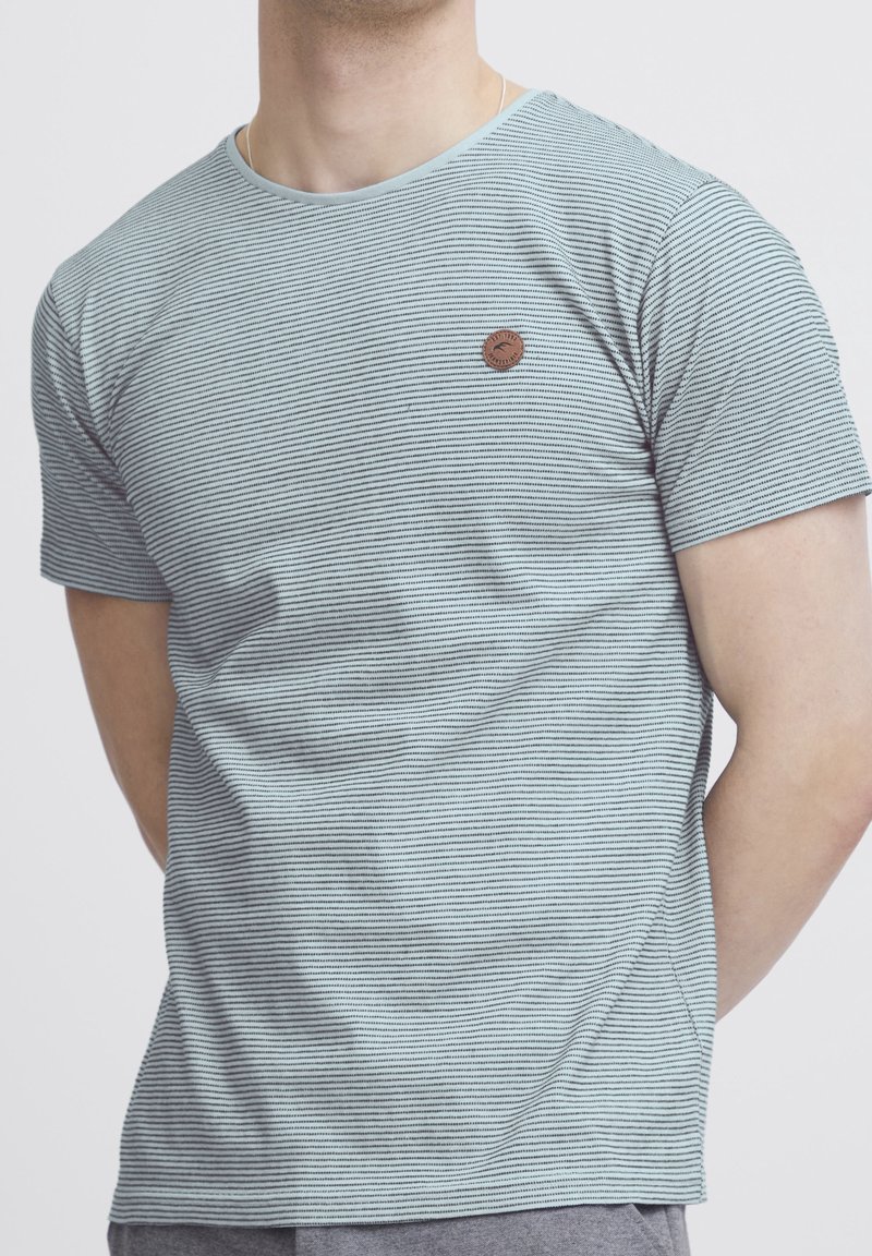 Homme portant un t-shirt à manches courtes à fines rayures bleu clair et blanches, avec un petit logo rond marron sur la poitrine gauche.
