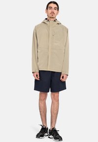 Chaqueta con capucha color beige con un patrón texturizado, cierre de cremallera y dos bolsillos frontales, combinada con pantalones cortos azul marino y zapatillas deportivas negras.