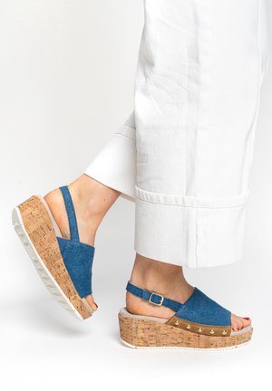Nohy v modrých džínových klínových sandálech s korkovou podrážkou a bílých kalhotách stočených nad kotníky, jedna noha mírně zdvižená.