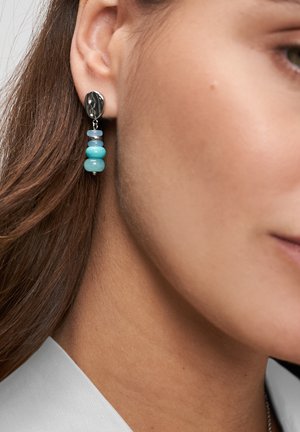 Zilveren en blauwe oorbel met kralen die aan het linker oor van een persoon met bruin haar en lichte huid hangt.