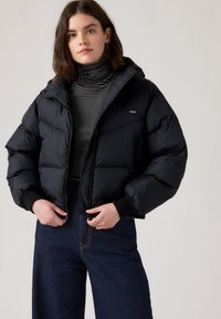 Casaco puffer preto com corte cropped, fecho de correr e capuz. Camisola de gola alta em mistura de algodão com riscas e calças de ganga escuras com costura visível.