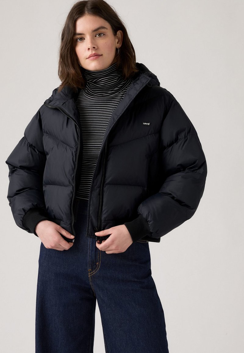 Casaco puffer preto com corte cropped, fecho de correr e capuz. Camisola de gola alta em mistura de algodão com riscas e calças de ganga escuras com costura visível.