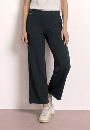 Femme portant un pantalon large noir, un haut sans manches blanc et des baskets blanches, debout contre un mur clair texturé.