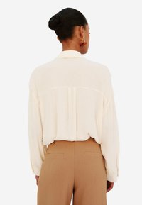 Blusa di leggera crema con colletto a camicia, vestibilità rilassata e maniche lunghe. Abbinata a pantaloni marroni su misura, con sottili tasche posteriori.