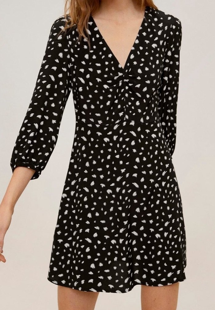 Robe noire avec des taches abstraites blanches, manches trois-quarts, encolure en V et détail froncé au niveau de la poitrine, portée par une femme.