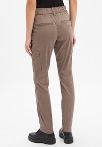 Pantalons cargo marron extensibles avec élastique à la taille, poches latérales et arrière, jambes fuselées et tissu doux et texturé. Bottes noires en dessous.