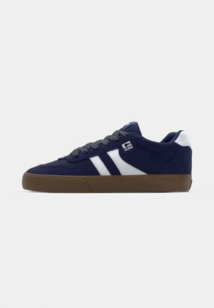 Navy blue suede sneaker with white stripes, grey laces, and a textured brown rubber sole. Features a logo on the side and a padded collar.